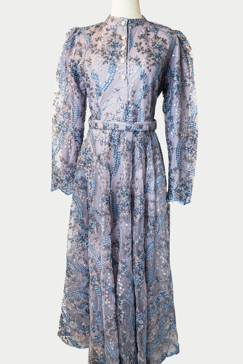 A stunning long dress in purple with sequins and delicate blue embroidery. The dress has mandarin collar, buttons, long sleeves, and is fully lined. There are hidden zips at the back and sleeves for ease of wear. The design is hijabi-friendly for modesty and style. It also includes a size-adjustable belt for a customized fit.