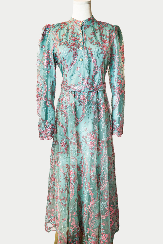 A stunning long dress in green with sequins and delicate pink embroidery. The dress has mandarin collar, buttons, long sleeves, and is fully lined. There are hidden zips at the back and sleeves for ease of wear. The design is hijabi-friendly for modesty and style. It also includes a size-adjustable belt for a customized fit.