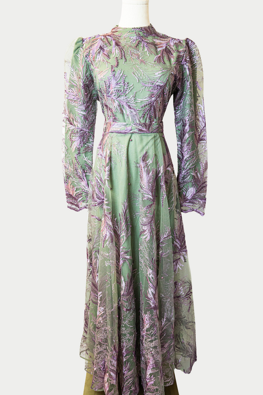 A stunning long dress in green with pearls and delicate purple embroidery. The dress has mandarin collar, long sleeves, and is fully lined. There are hidden zips at the back and sleeves for ease of wear. The design is hijabi-friendly for modesty and style. It also includes a size-adjustable belt for a customized fit.