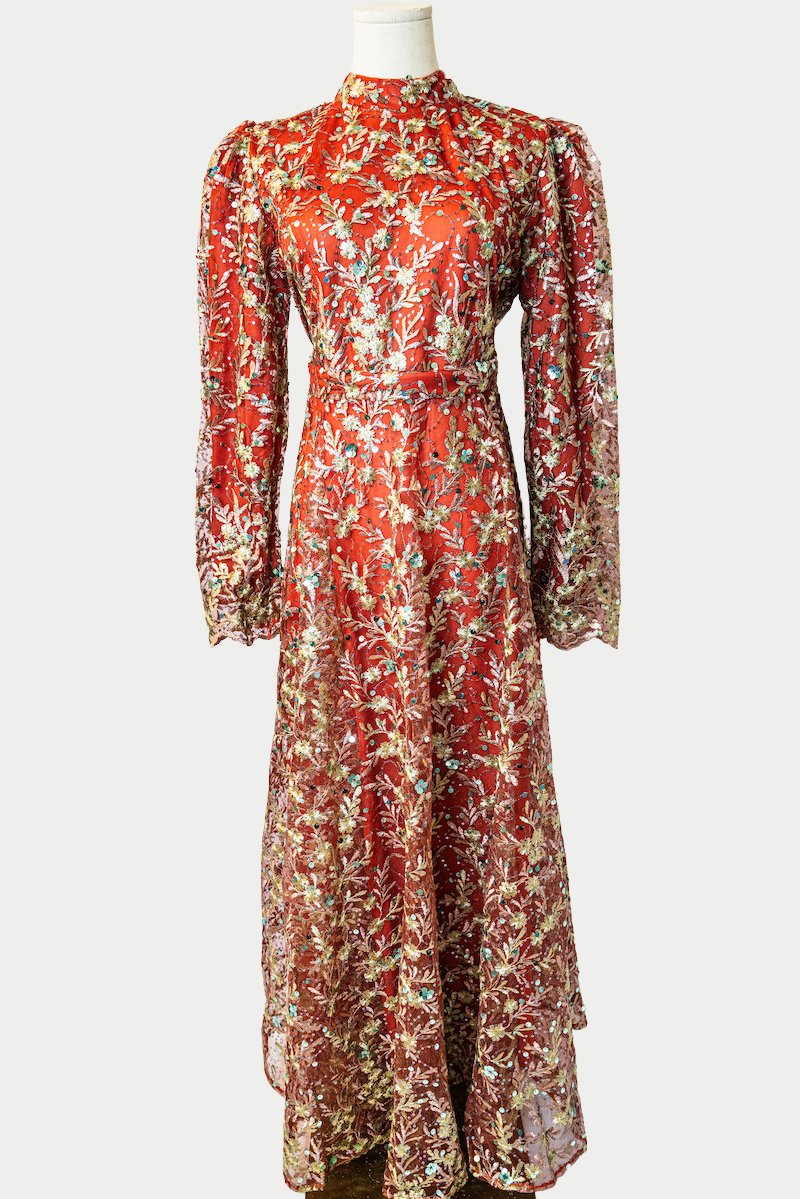 A stunning long dress in orange with sequins and delicate golden embroidery. The dress has mandarin collar, long sleeves, and is fully lined. There are hidden zips at the back and sleeves for ease of wear. The design is hijabi-friendly for modesty and style. It also includes a size-adjustable belt for a customized fit.